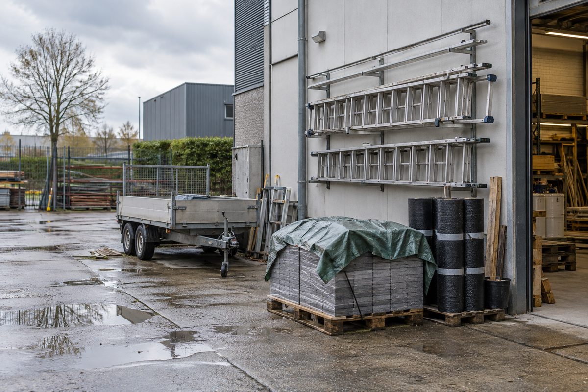 Arbejdsplads med stiger på væggen, en trailer og paller med tagmaterialer efter regn